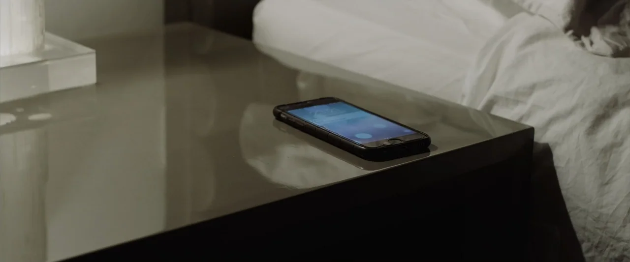 The Thorn On His Side (2014) movie still: closeup — A modern nightstand surface holds a smartphone showing a screen notification in the dark. The…