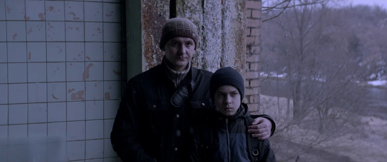Loveless (2017) movie still: medium — A man and a boy stand inside a partially constructed, weathered brick structure. The background features a…