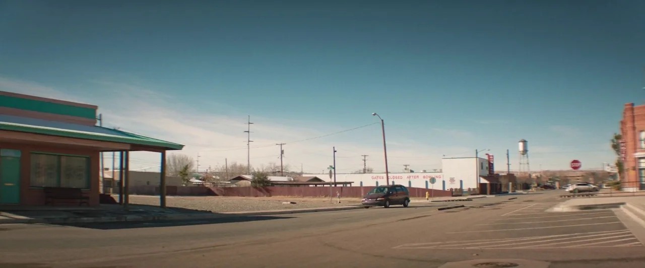 The Long Dumb Road (2018) movie still: establishing — A sparsely populated town street features a low-slung building with a covered walkway. A small…