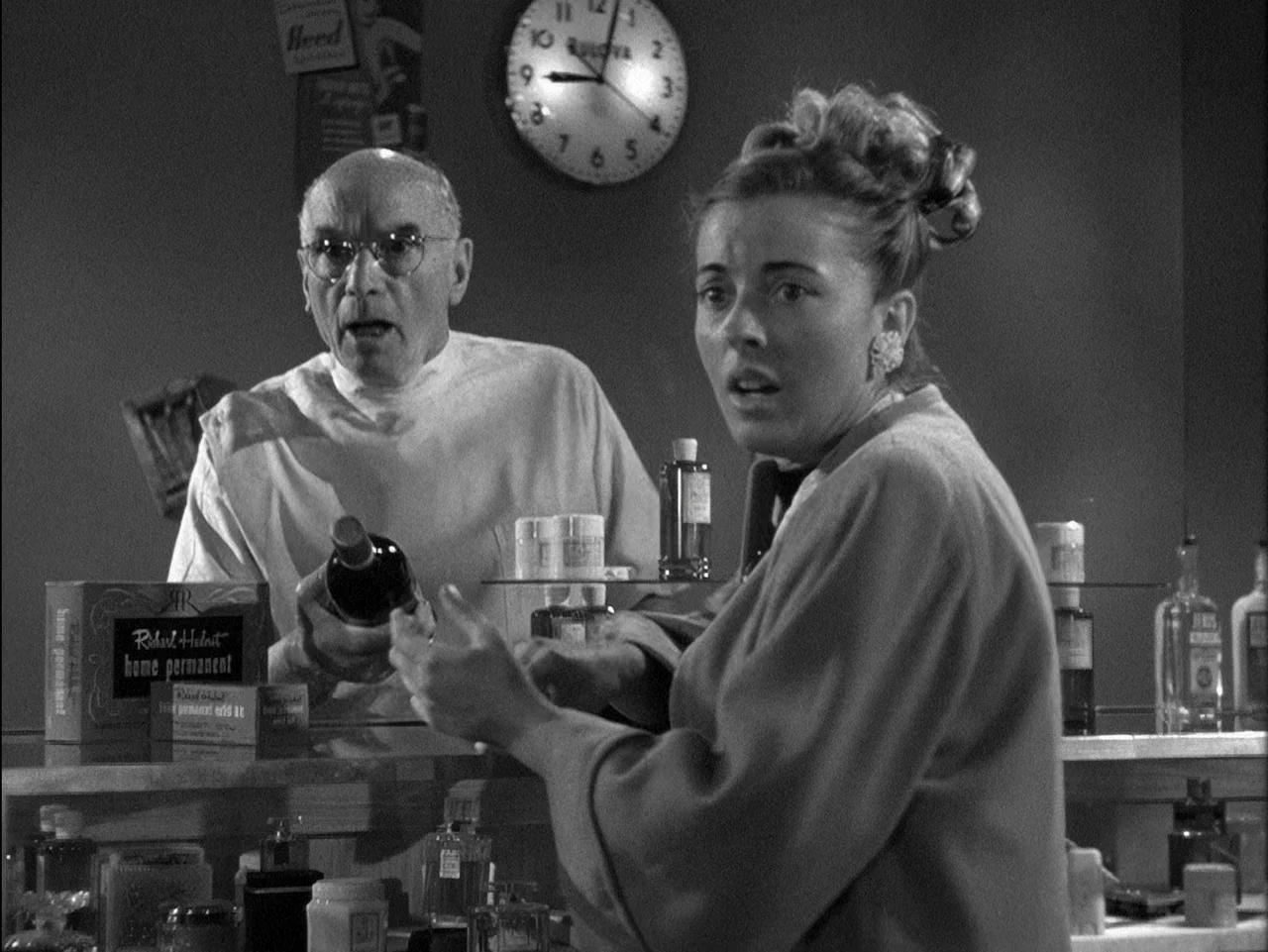 D.O.A. (1949) movie still: medium — A 1950s drugstore counter displays various beauty products and glass bottles. An elderly man and a woman stand…