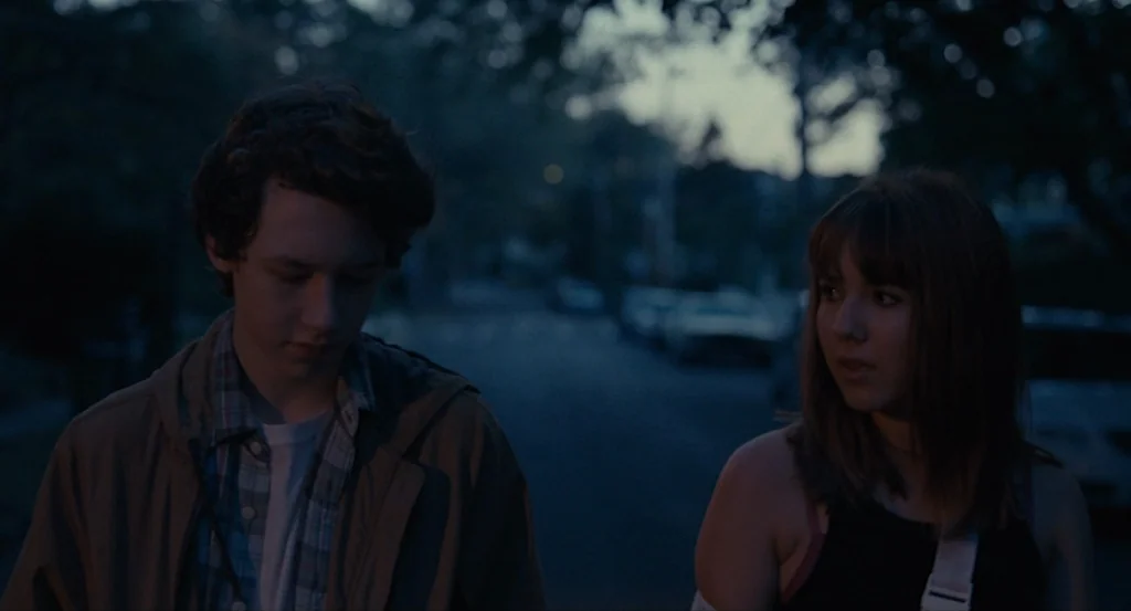 Louder Than Bombs (2015) movie still: medium — A suburban residential street at night with parked cars visible in the background blur. A teenage boy…