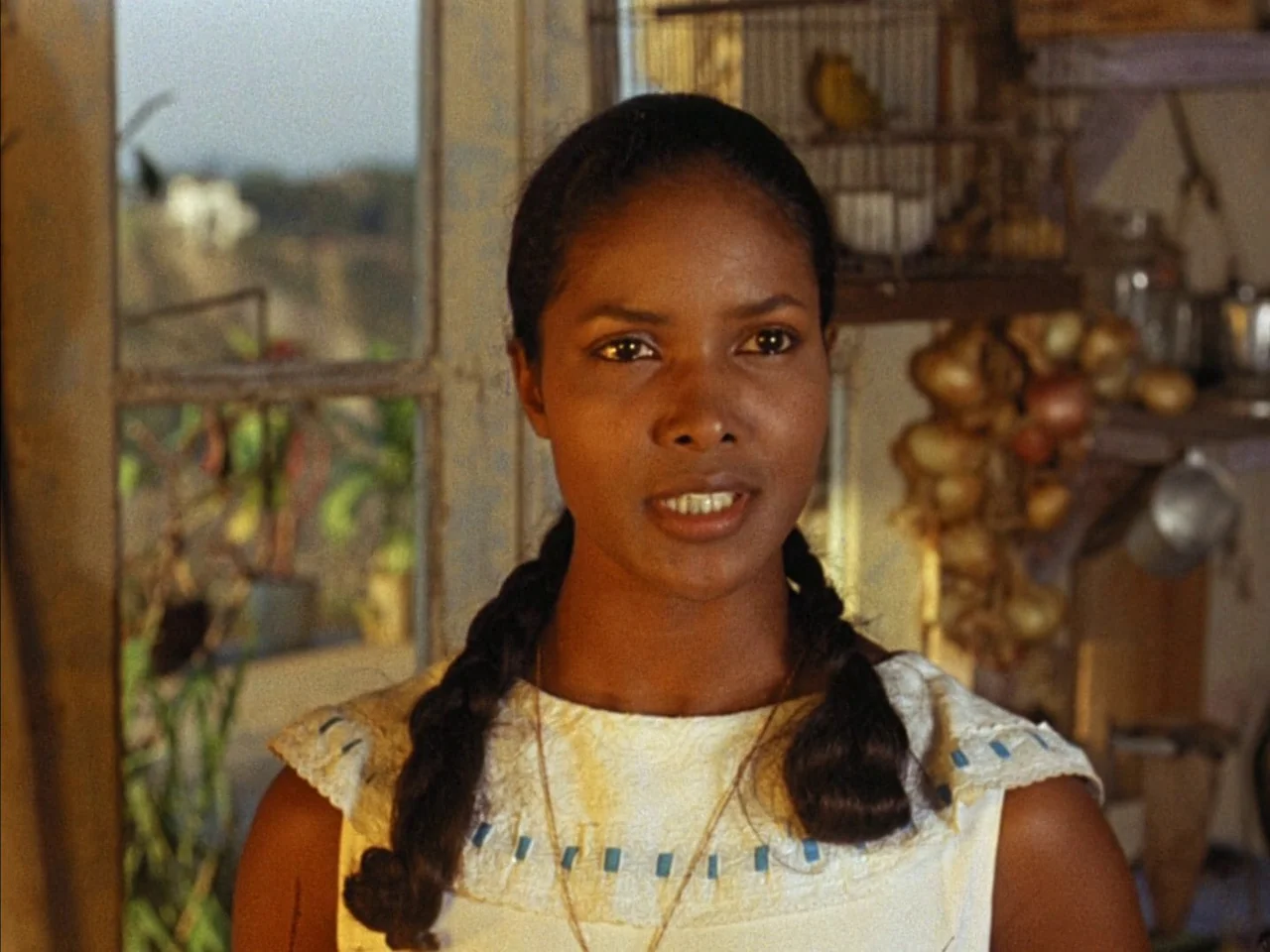 Black Orpheus (1959) movie still: closeup — A rustic cottage interior shows a window looking out onto a blurred bright exterior. A young woman in a…
