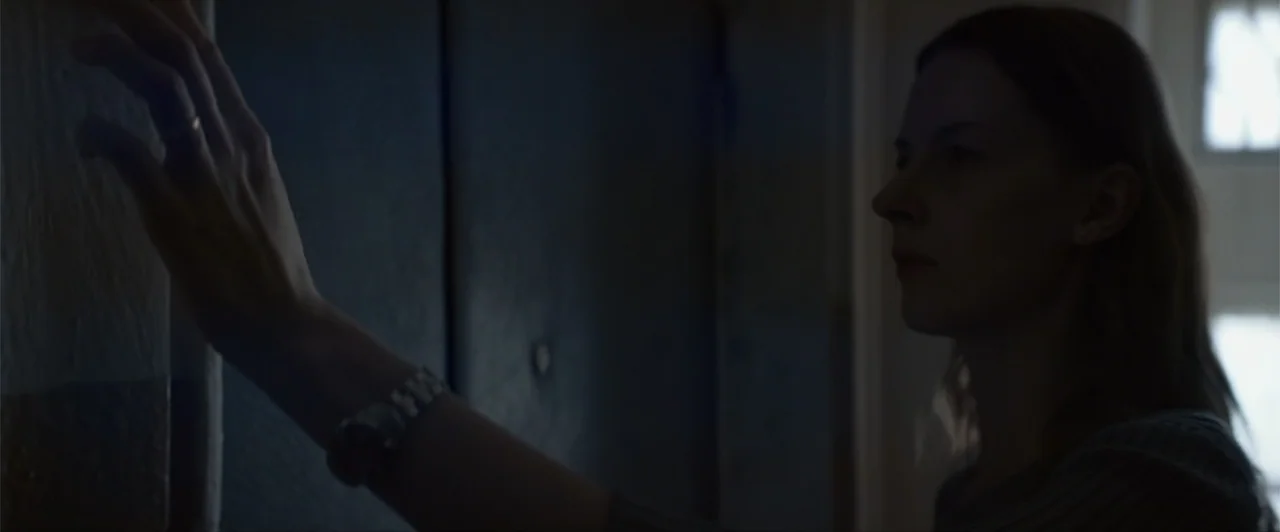 La herencia (2010) movie still: medium — An interior hallway features dark walls and a glimpse of a bright rectangular window. A young woman in a…