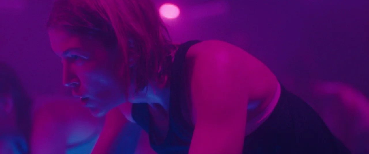 I Care A Lot (2020) movie still: closeup — A dimly lit room filled with neon purple and magenta light. A woman in athletic wear cycles intensely on…