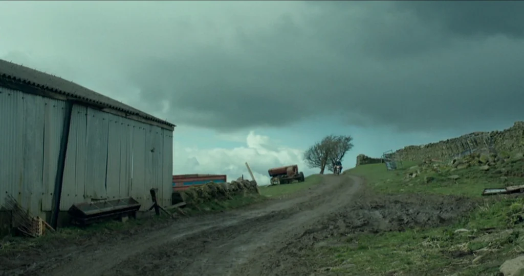God’s Own Country (2017) movie still: wide — A rural landscape features a corrugated metal structure beside a winding muddy dirt road. A distant…