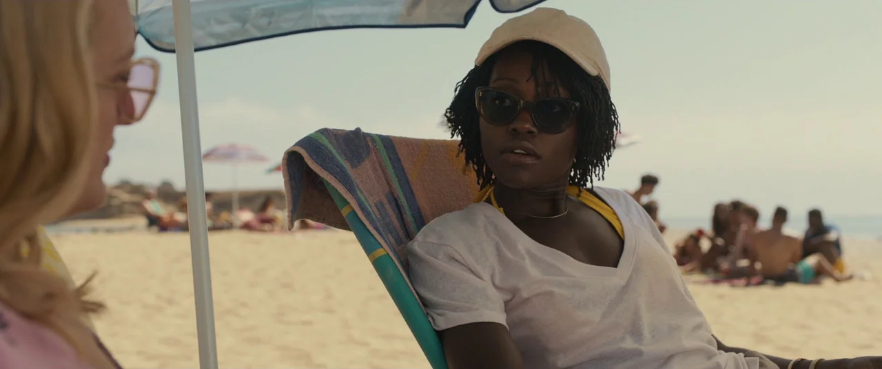Us (2019) movie still: medium — A sunny sand beach with people lounging in the distance. One woman sits in a folding chair beneath a blue umbrella…