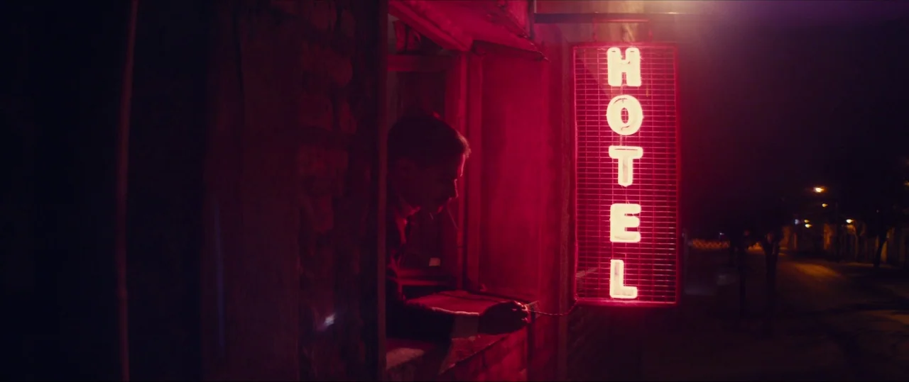 Neruda (2016) movie still: medium — A man peers out from a window adjacent to a glowing red hotel sign. The dark facade of the building is…