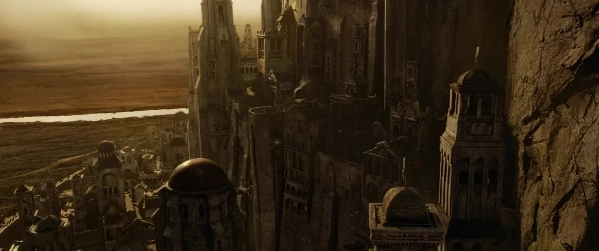 A vast fantasy stone city is built directly into the side of a sheer cliff. Below the structures, a river winds through the flat, golden landscape of the valley.