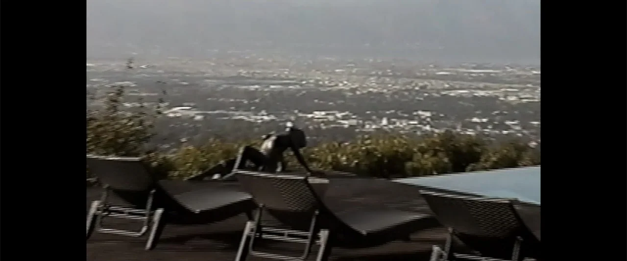 A Scam Called Love (2025) movie still: wide — A hillside deck overlooks a vast sprawling city under bright daylight. A lone woman sits on a lounge…