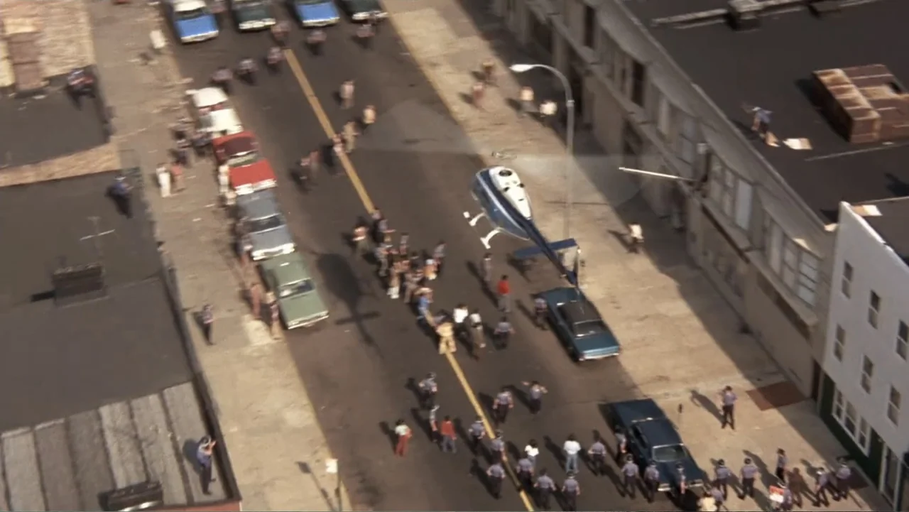 Dog Day Afternoon (1975) movie still: wide — A dense urban street with parked vintage automobiles and a crowd of people below. A blue and white…