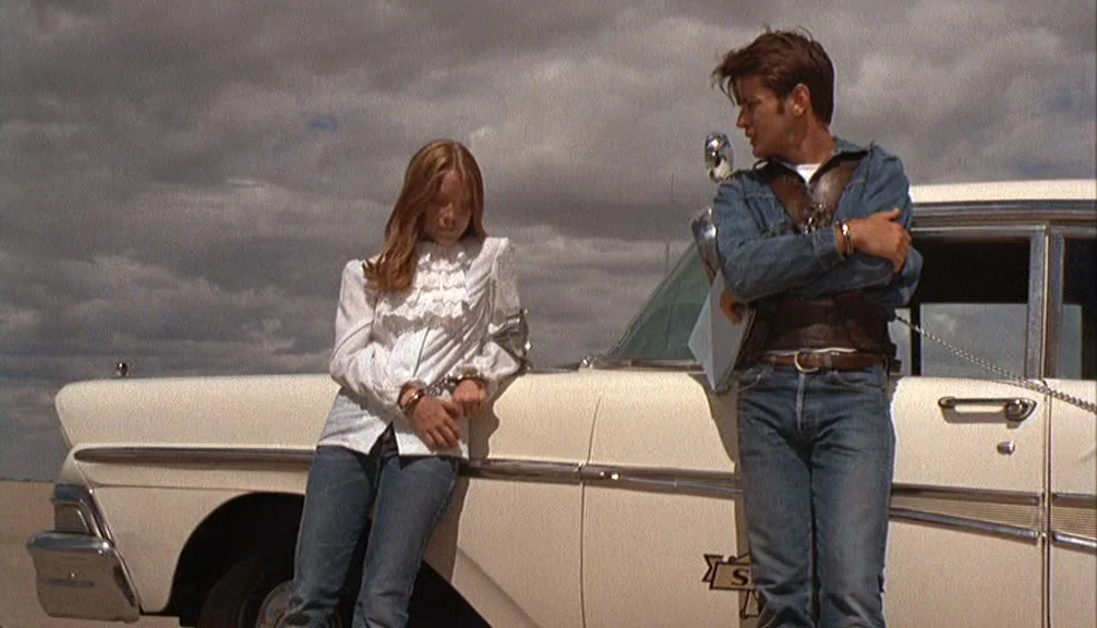 Badlands (1973) movie still: fullbody — Two individuals stand beside a pale sedan against a backdrop of a cloudy sky. The male leans against the…