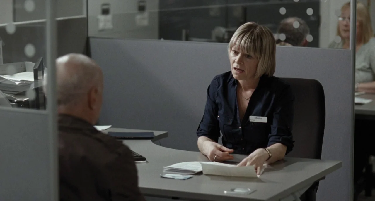 I, Daniel Blake (2016) movie still: medium — An office cubicle features gray partitions and a desk with scattered administrative papers. A woman…