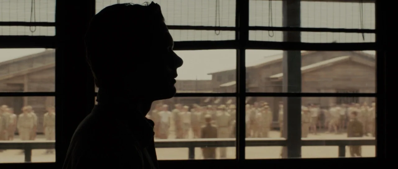 Unbroken (2014) movie still: medium — A dark interior room features a window overlooking an open dirt training yard. A man stands in silhouette by…