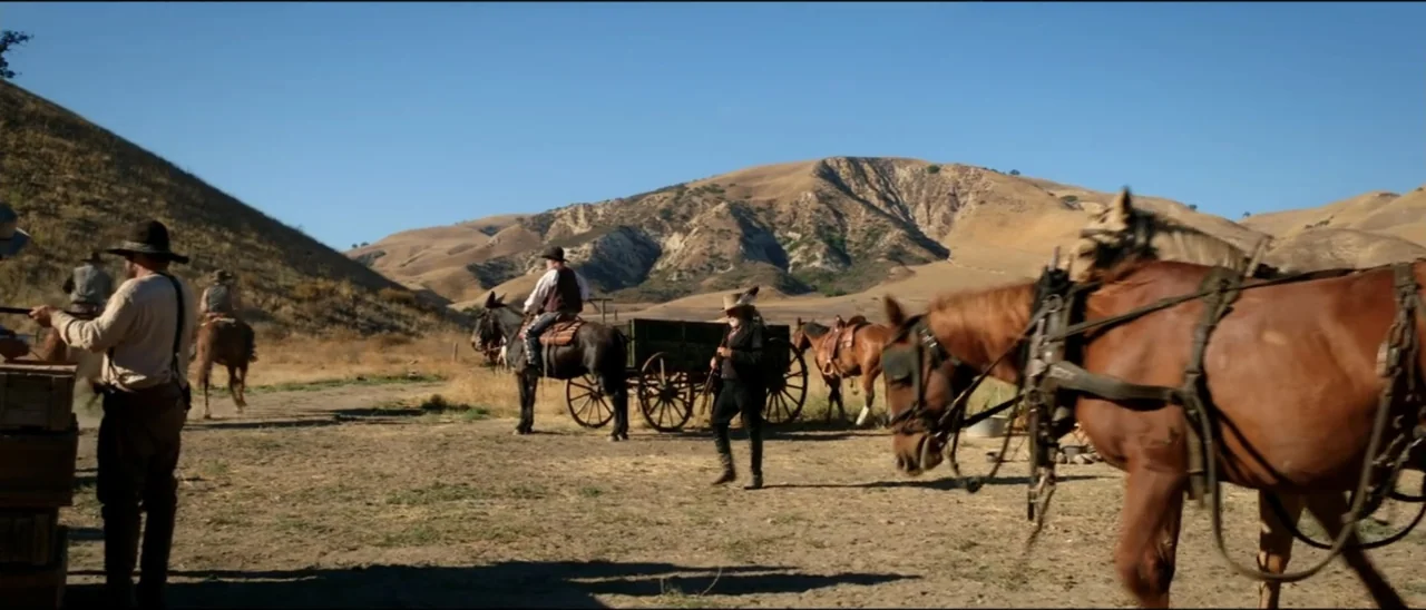 Dead for a Dollar (2022) movie still: wide — An arid, sun-drenched lot sits beneath rolling dry hills under a clear blue sky. Men in period western…