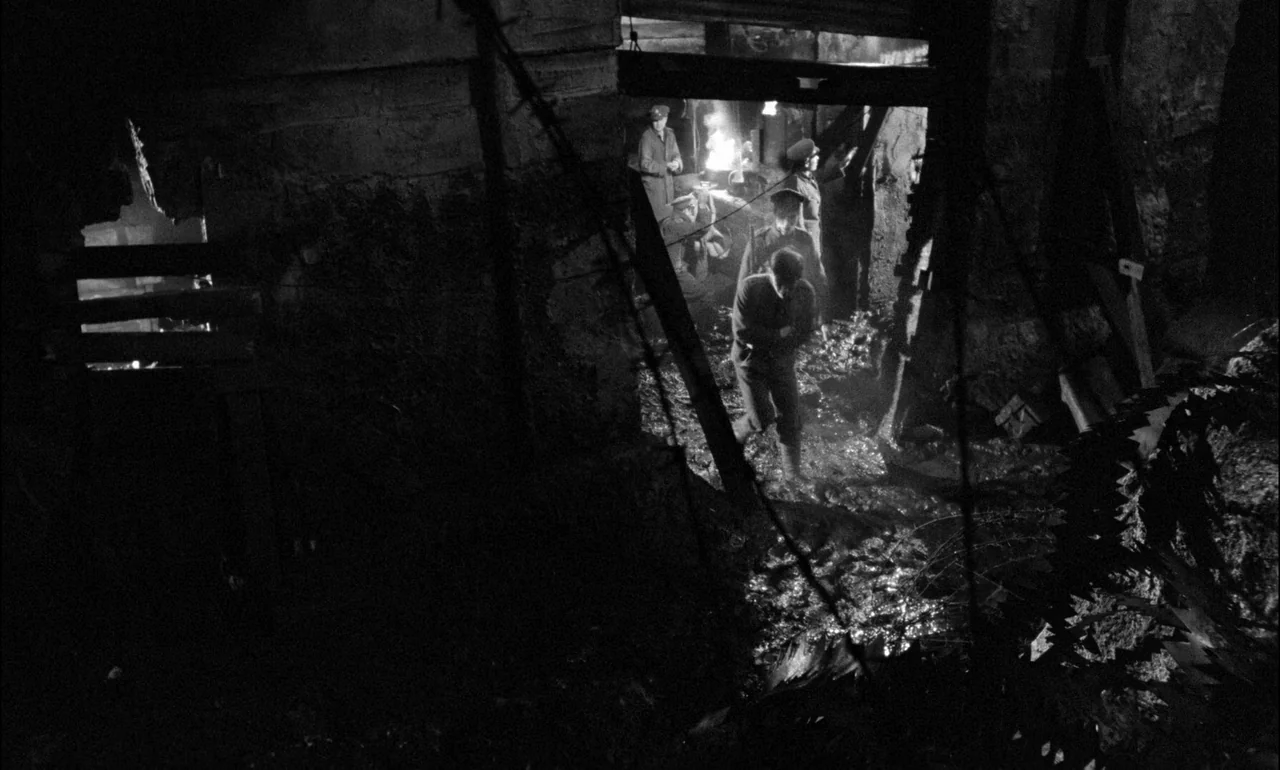 King & Country (1964) movie still: wide — A dark muddy trench with barbed wire silhouettes in the foreground and sides. Several soldiers cluster…