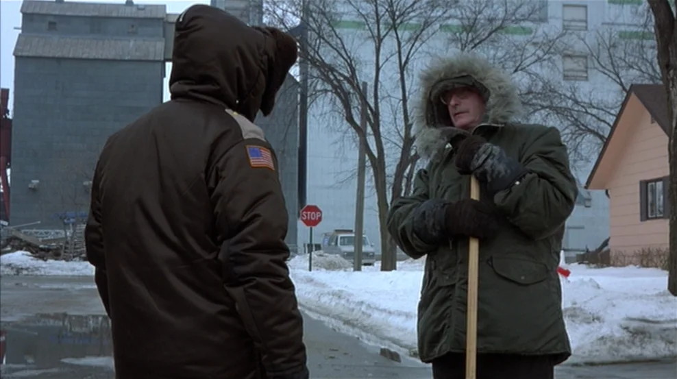 Fargo (1996) movie still: medium — An overcast winter street scene features a residential area with snow-covered ground. Two men in heavy winter…