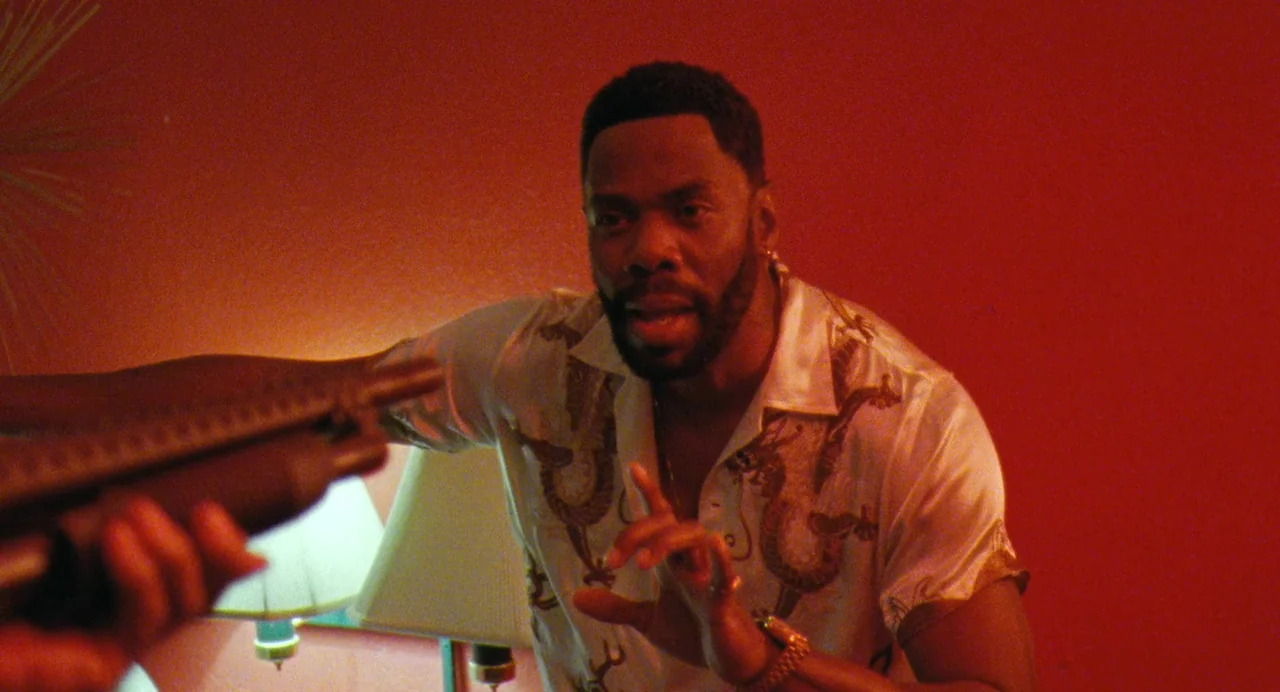 Zola (2020) movie still: medium — A dimly lit room with vibrant red walls and a lamp on the table. A man stands wearing a patterned silk shirt with…