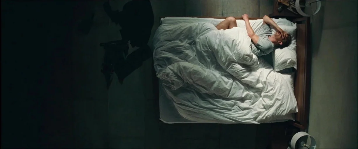 Layer Cake (2004) movie still: wide — A minimalist room contains a bed placed on a dark, polished floor surface. A man lies under a white duvet with…