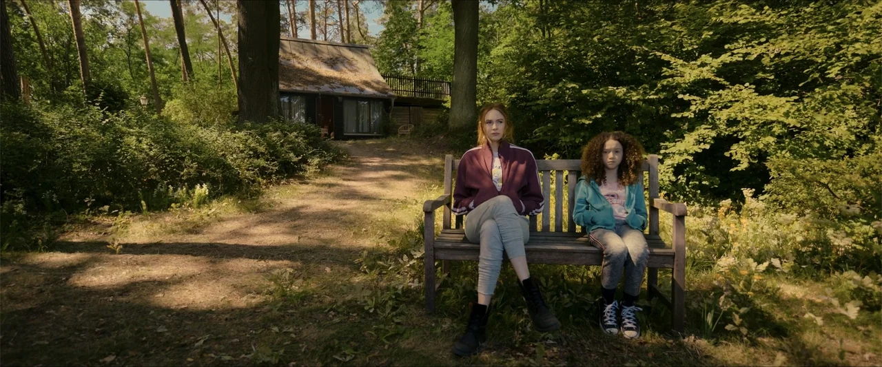 Gunpowder Milkshake (2021) movie still: fullbody — Two individuals sit on a wooden bench situated within a dense wooded area. A young woman and a…