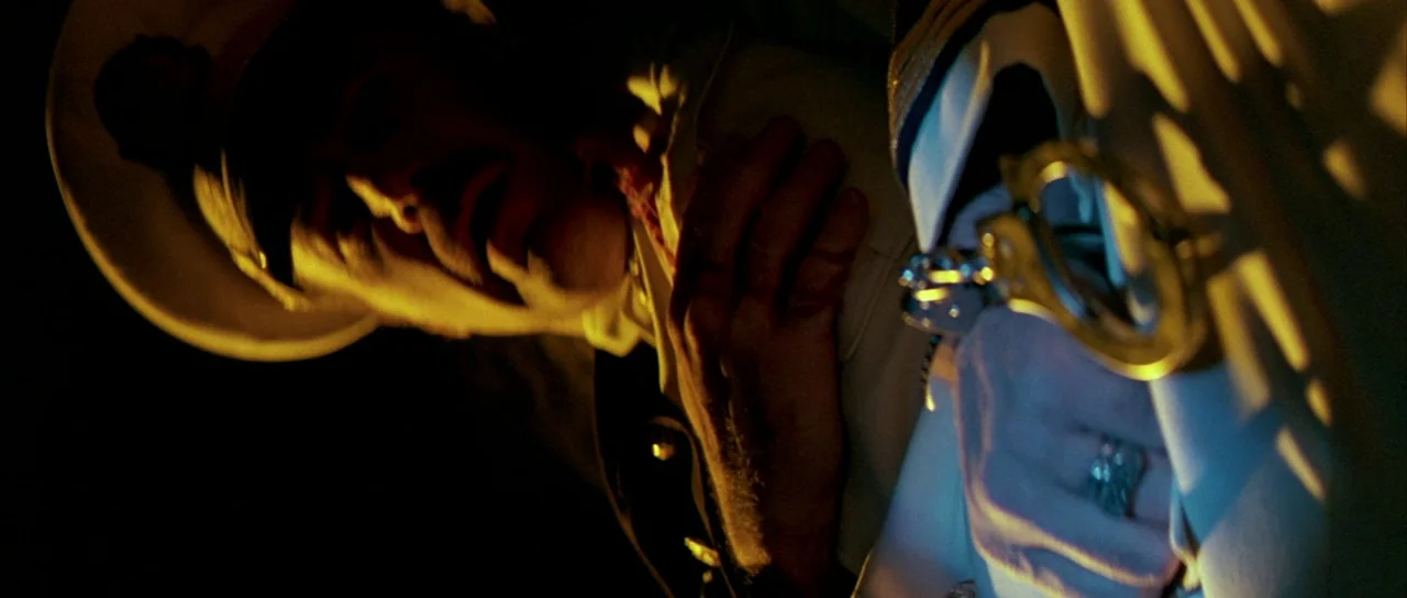 Querelle (1982) movie still: closeup — A dark, cramped interior lit by harsh, low-angle yellow light. A man in uniform grips his chest while…