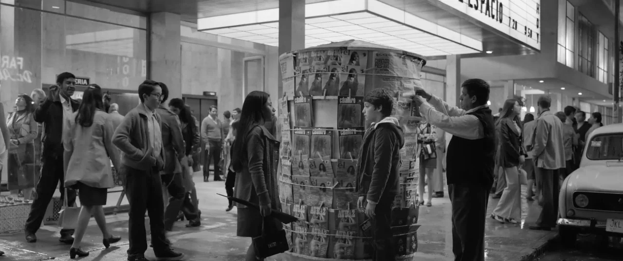 Roma (2018) movie still: wide — A bustling city street outside a movie theater at night with pedestrians walking by. A woman and a young boy stand…