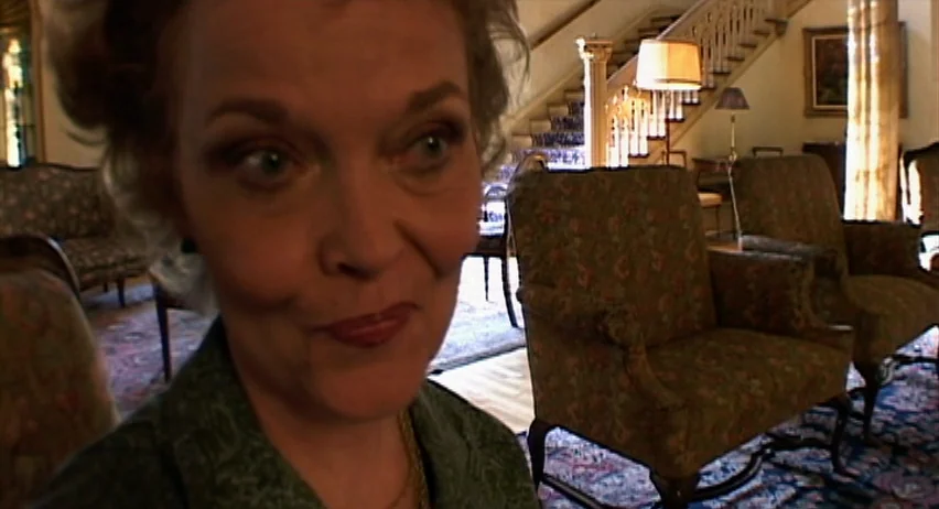 Inland Empire (2006) movie still: closeup — A grand foyer with patterned carpets, upholstered armchairs, and a staircase in the background. A woman…