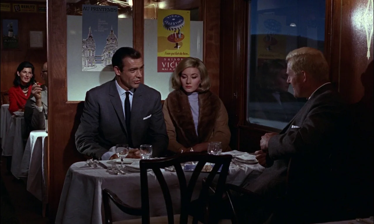 From Russia With Love (1963) movie still: medium — A wood-paneled dining car on a train features vintage advertisements on the wall. Three people…