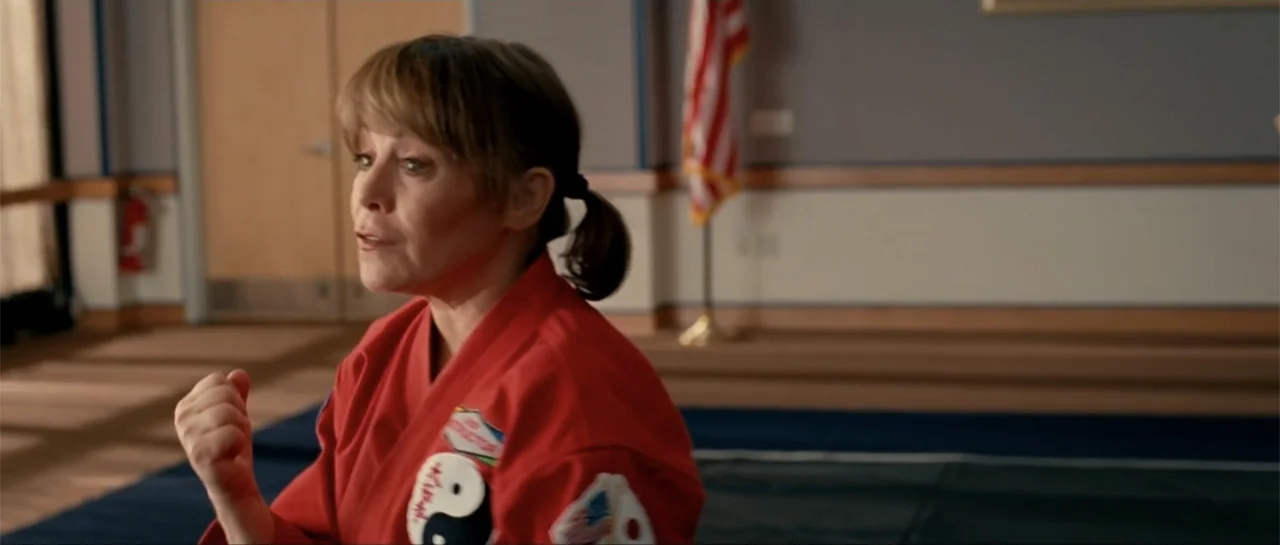 The Rebound (2009) movie still: medium — A woman stands in a martial arts studio with an American flag behind her. She wears a red martial arts…