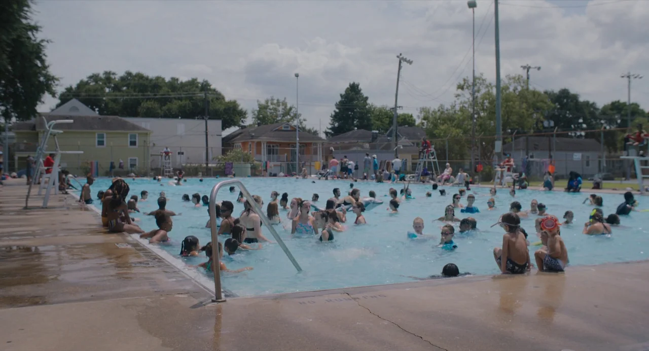 Causeway movie still: wide — A crowded public swimming pool filled with numerous people occupies the center frame. Suburban houses and utility poles…
