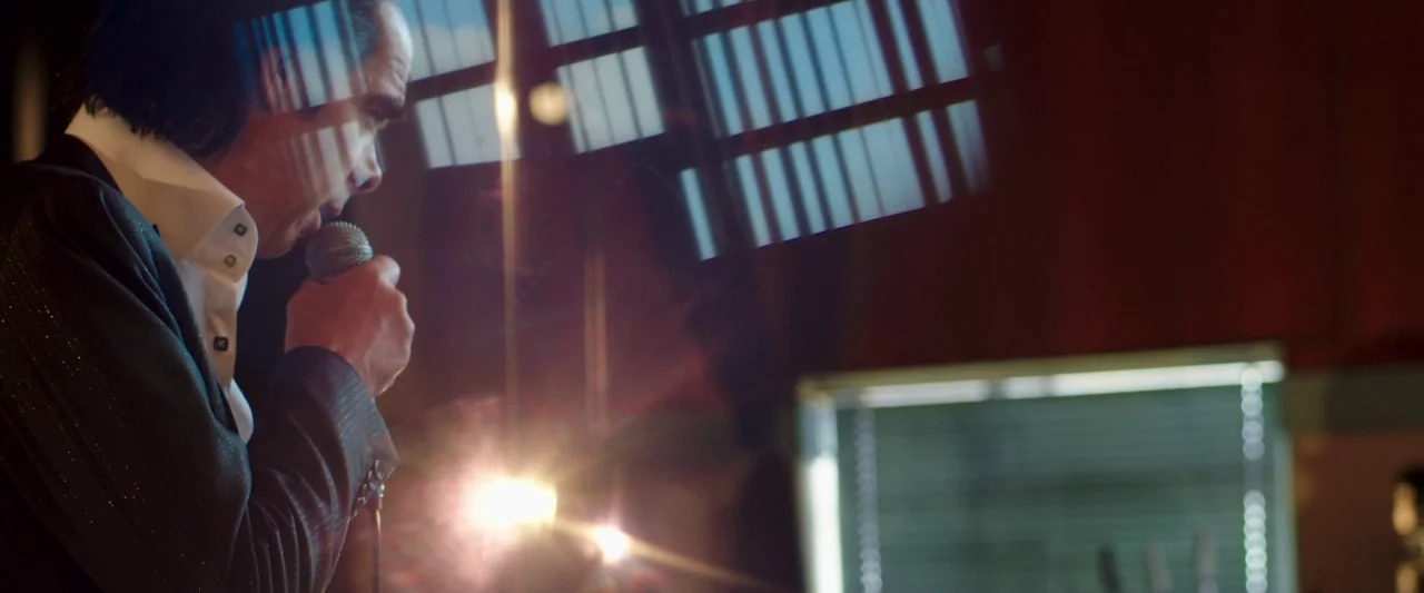 20,000 Days on Earth (2014) movie still: medium — A dark recording studio with dark wood paneling and a blurred window with blinds. A man in a dark…