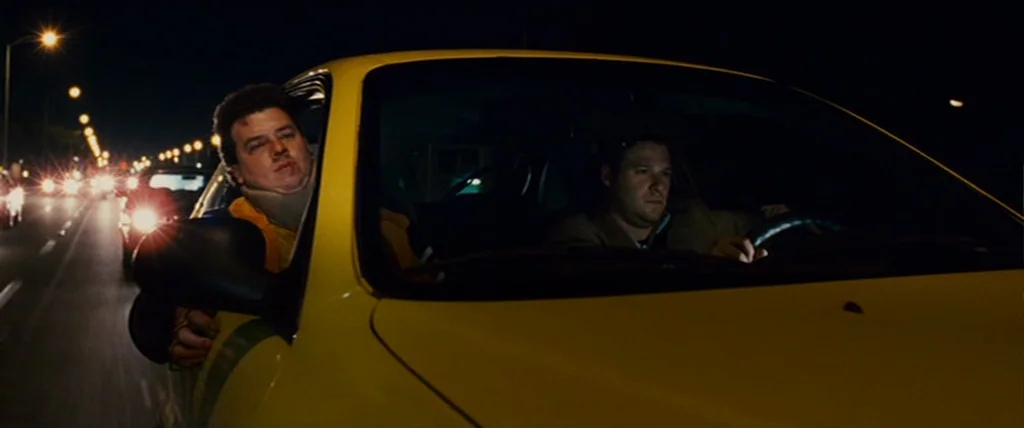Pineapple Express (2008) movie still: medium — A yellow vehicle travels along a city street at night under rows of lamps. Two men sit in the front…