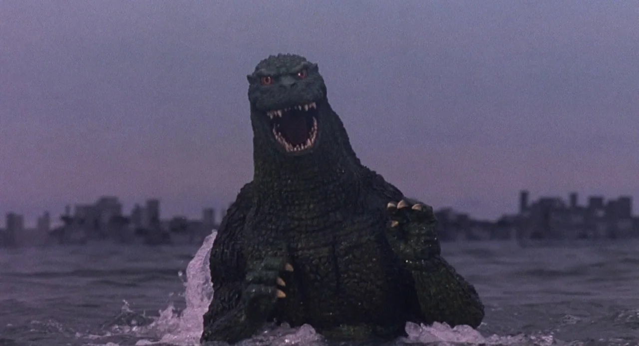 Godzilla vs. Destoroyah (1995) movie still: medium — Godzilla stands in dark ocean water against a distant, blurry city silhouette at twilight. The…