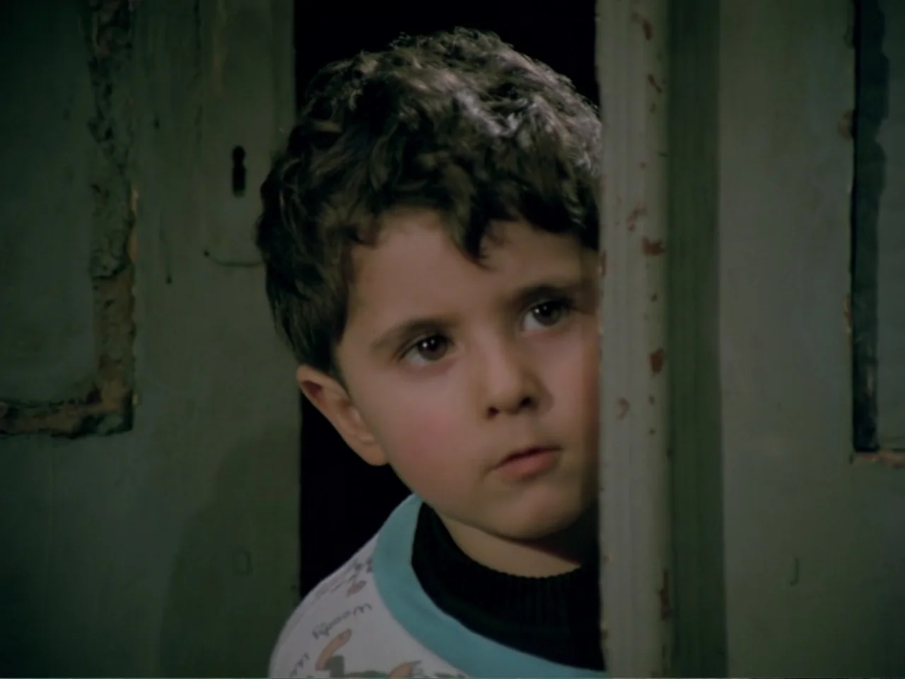 Don't Let Them Shoot the Kite (1989) movie still: closeup — A young boy peers from behind a slightly open, peeling light-green painted door. He…