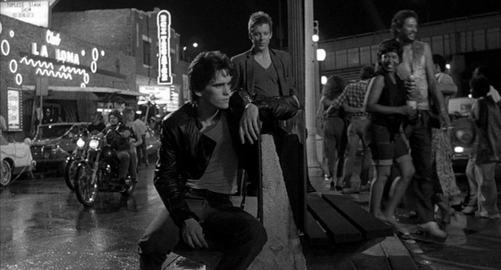 Rumble Fish (1983) movie still: medium — A busy urban sidewalk at night features glowing neon signs for local businesses. A young man sits on a…