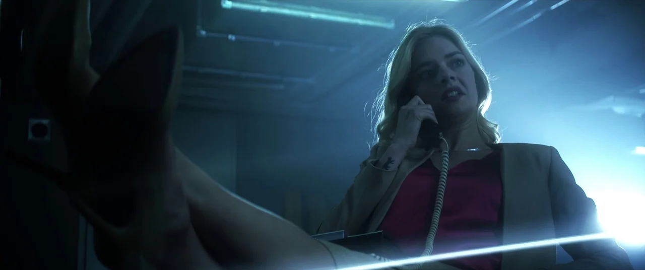 Mayhem (2017) movie still: medium — A dimly lit room with overhead lights and a desk in the foreground. A blonde woman sits at the desk with her…