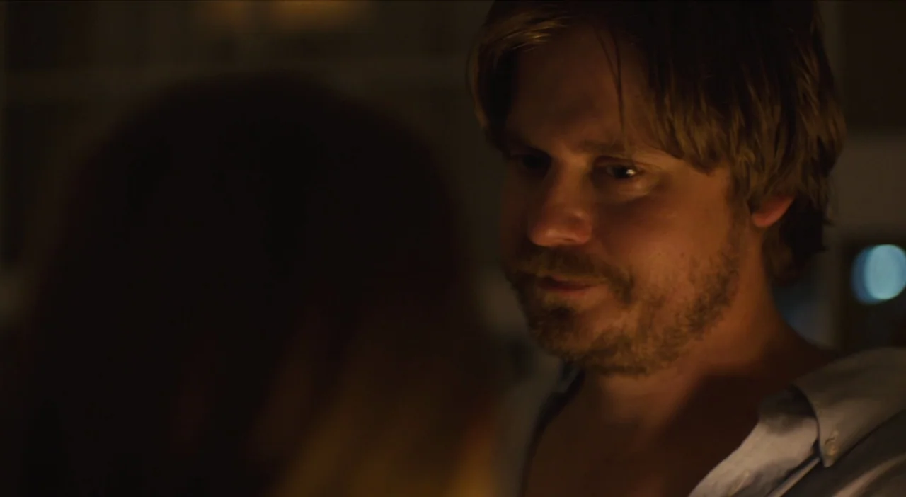 The Comedy (2012) movie still: closeup — A dark indoor space with a man positioned in the foreground right. The man turns his head slightly while…