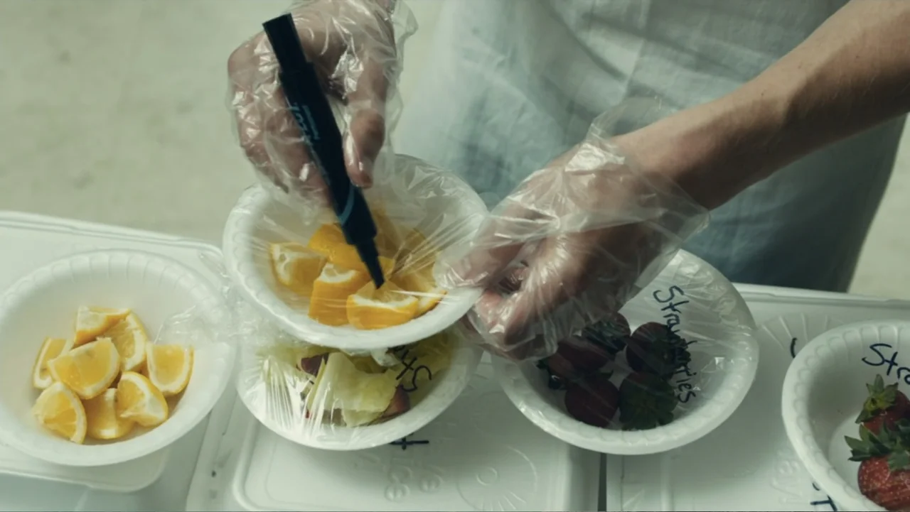 Camp X-Ray (2014) movie still: closeup — A white sterile workstation contains several styrofoam bowls covered in clear plastic wrap. Hands wearing…