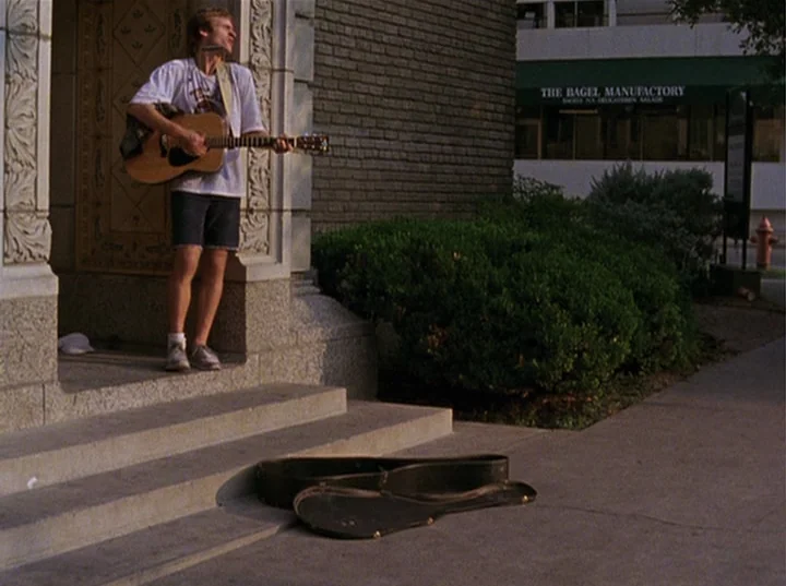 Slacker (1991) movie still: fullbody — A man stands playing a guitar in the entrance of a brick building. An empty instrument case rests on the…