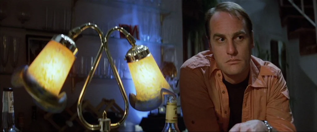 Poltergeist movie still: medium — A dimly lit indoor space features a brass lamp with two warm, glowing shades. A man sits at a surface, looking off…