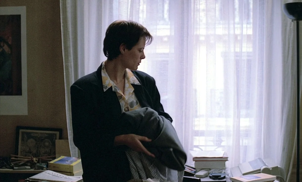 A Tale of Springtime (1990) movie still: medium — A room with a desk cluttered with papers and books before a large window. A woman wearing a navy…