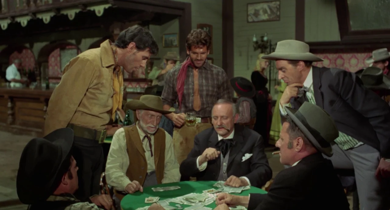 Johnny Colt (1966) movie still: medium — A crowded 19th-century frontier saloon with wooden walls and poker tables. Six men sit and stand around a…