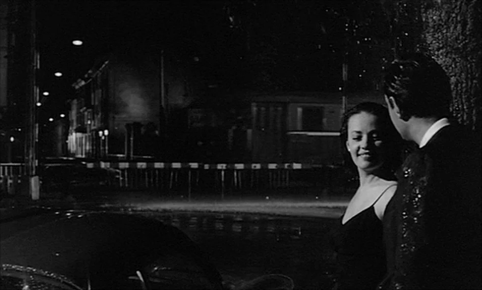 La Notte (1961) movie still: medium — A dark, quiet road at night features a parked vehicle and distant streetlights. A man and a woman stand…