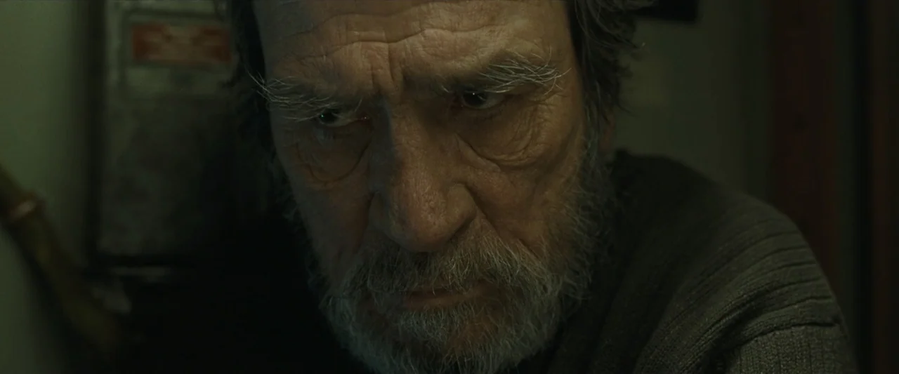 Ad Astra (2019) movie still: closeup — A dimly lit interior space with a man in the foreground. An elderly man with a grey beard stares downwards…