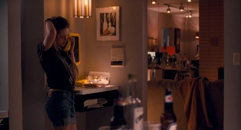 Adventureland (2009) movie still: medium — A dimly lit alcove in a living space features a wall-mounted phone and framed artwork. A woman stands in…