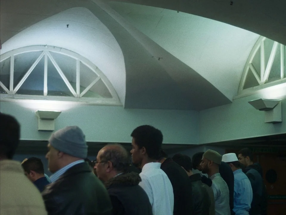 Mogul Mowgli (2020) movie still: medium — A group of men stand in a row within a building featuring vaulted ceilings. They are oriented in the same…