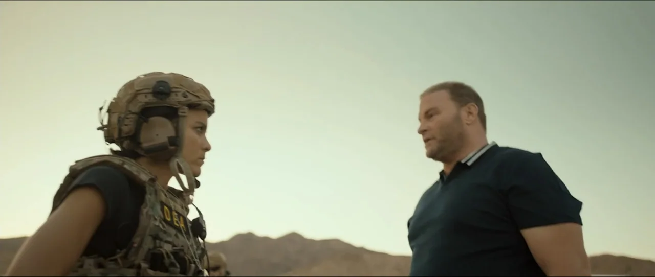 American Sicario (2021) movie still: medium — A vast, barren desert landscape stretches out behind two people standing near each other. A soldier in…