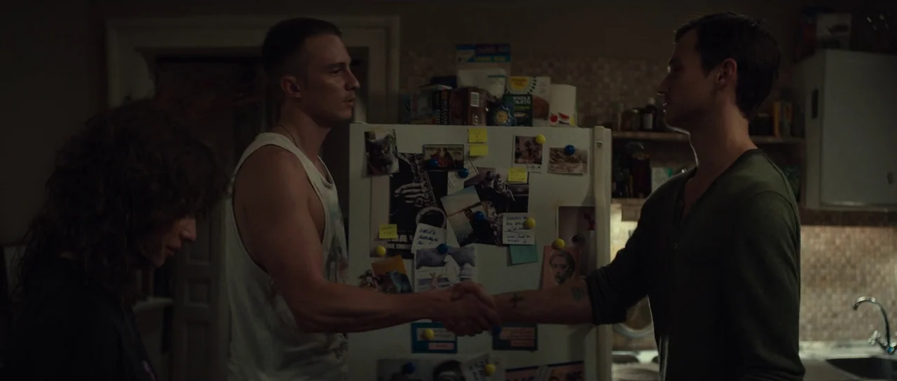 Hellraiser (2022) movie still: medium — An interior kitchen setting features a refrigerator covered with numerous photographs and notes. Two men…