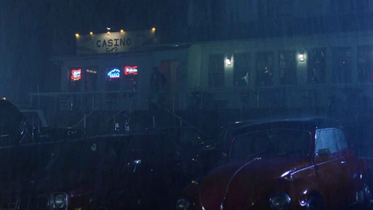 Friday the 13th Part 2 (1981) movie still: wide — A dark, rainy parking lot at night sits in front of a building. The building displays illuminated…