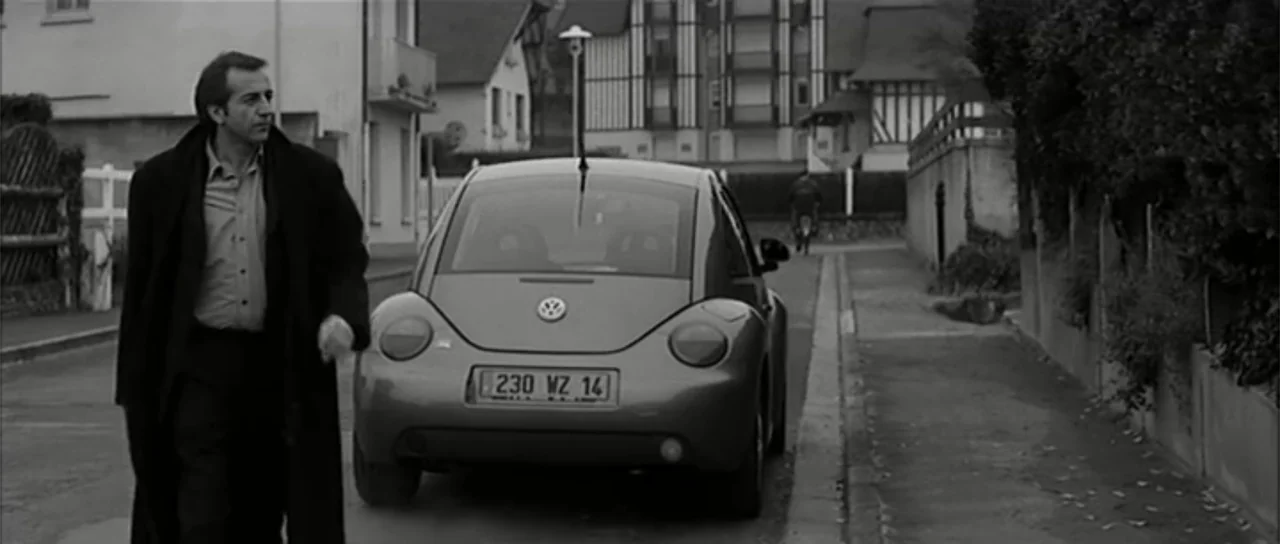 13 Tzameti (2006) movie still: fullbody — A suburban street scene features residential houses and a parked Volkswagen Beetle car. A man walks in the…