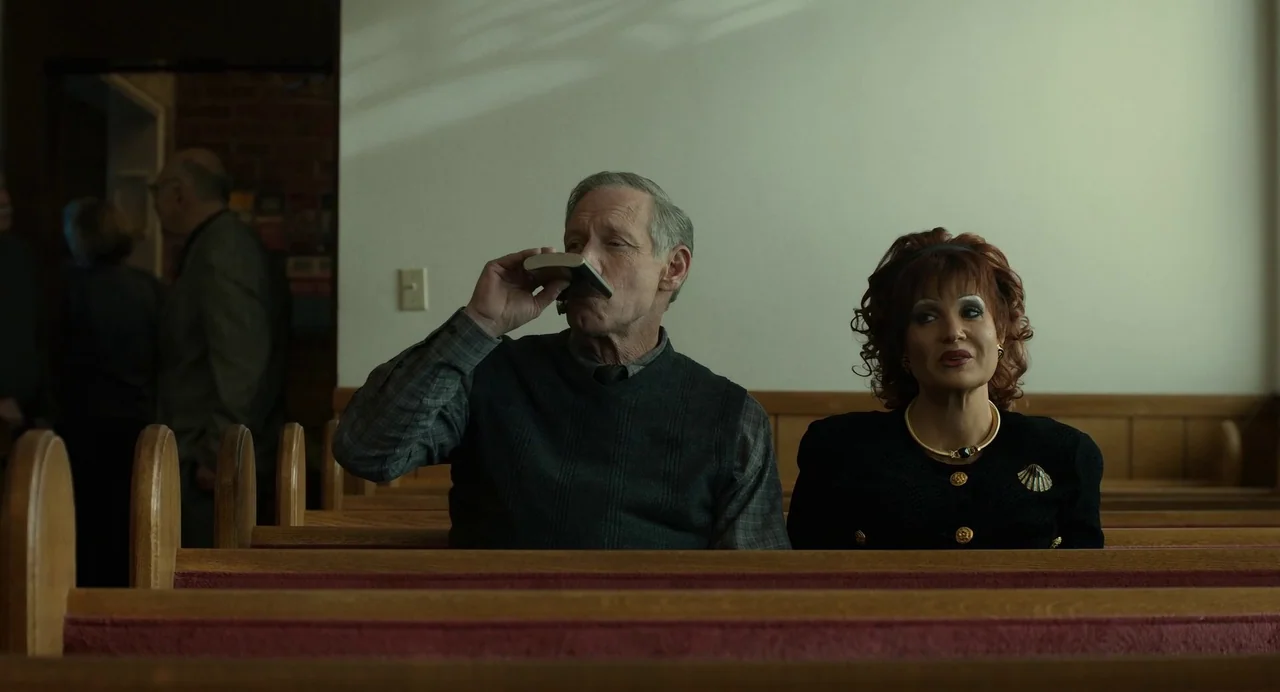 The Eyes of Tammy Faye (2021) movie still: medium — An interior space with rows of wooden benches facing toward the front. Two individuals sit…